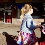 Photo of a young child with blonde hair wrapped up in a ponytail and wearing a floral dress, facing away. She pushes a pram in front of a shop named ‘Korona’.