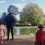 Photo of an adult, with two children standing behind her – one with bunches, one with plaited braids. They face away, staring at the lake and the trees.
