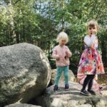 Photo of two children in pink, having climbed up boulders in woodland. One holds at a lolly, the other looks to a parent.